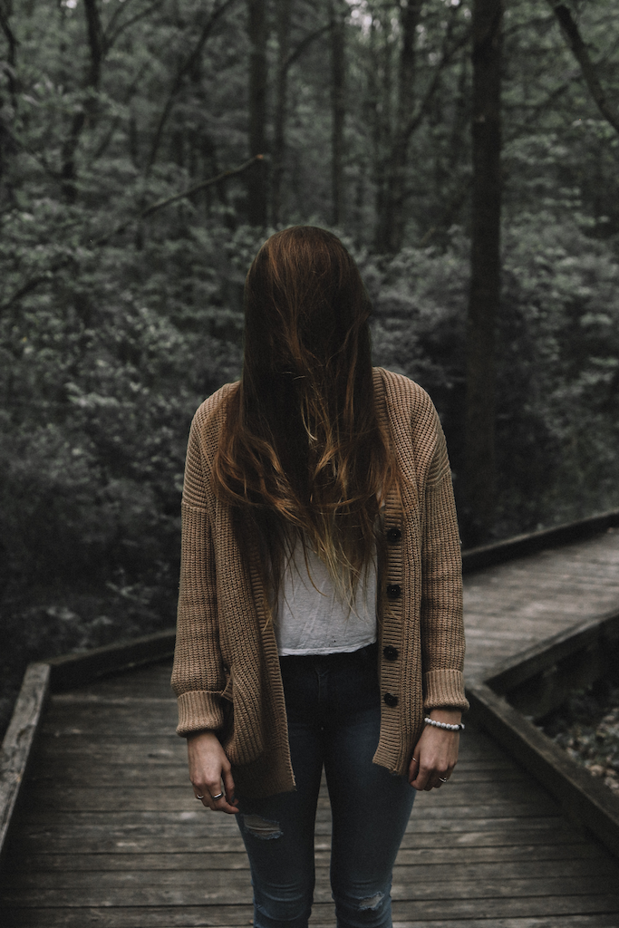 A woman hides her face in her hair while wearing a tan-colored Oasis cardigan in the woods.