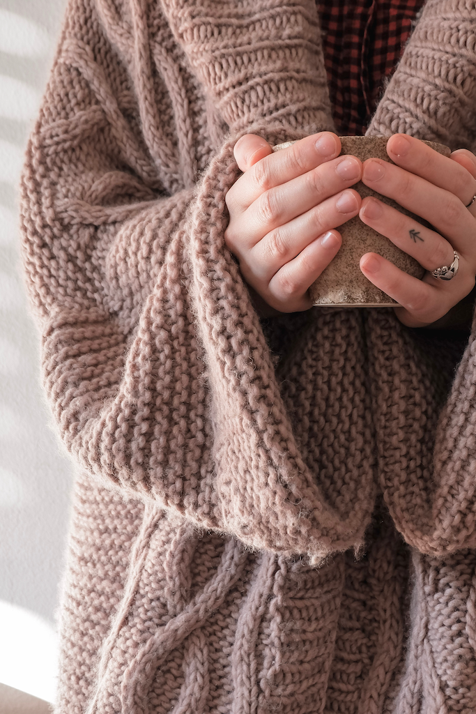 A woman drinks a cup of coffee in her brown, waterfall-styled Oasis womens cardigan.