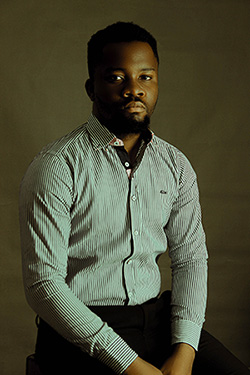 A man sits dressed in a striped green Oasis dress shirt and black Oasis slacks.