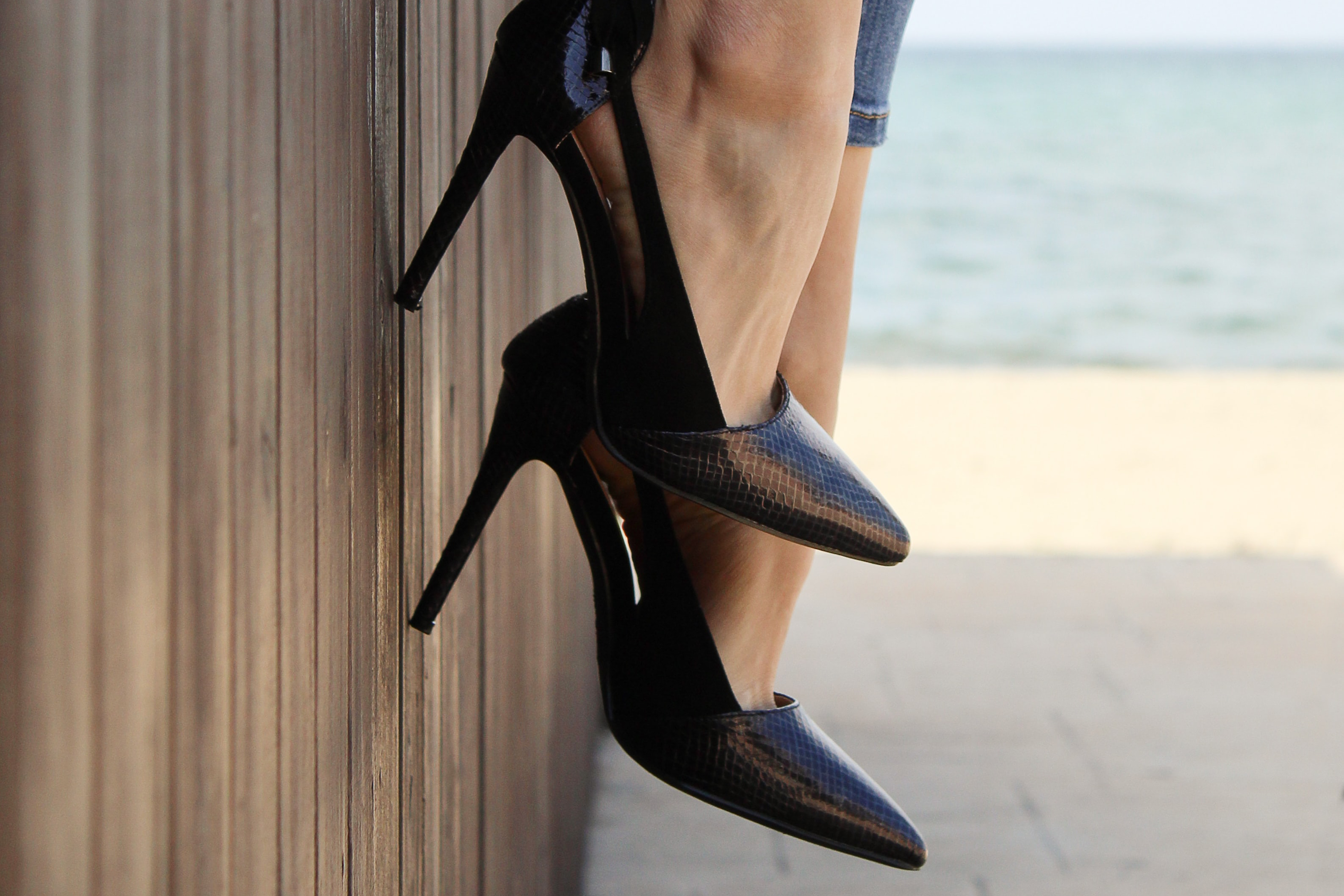 A woman sits beachside and dangles her feet while wearing a pair matte black Oasis heels.