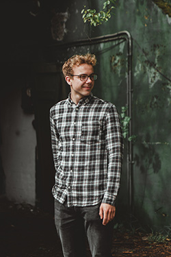 A man wearing a black-white Oasis men's plaid shirt stands near a forest-wall backdrop.